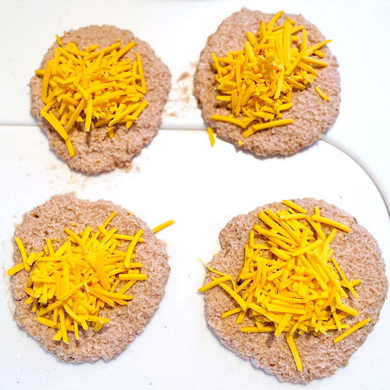 Divide la carne vegetal SoMeat en 4 partes iguales. Dale a cada parte forma de hamburguesa y pon por encima de cada una de ellas el queso rallado vegano. Luego, fríelas a fuego medio hasta que queden doradas.